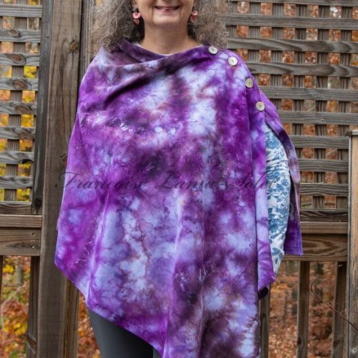 “Handmade ice-dyed poncho wrap in shades of purple, lavender, gray, and khaki, worn by a smiling woman outdoors.”